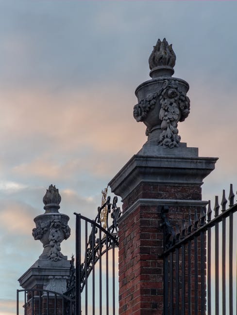A historic wrought iron gate with ornate finials and decorative stone urns atop brick pillars, set against a partly cloudy sky during either dawn or dusk. The gate's black ironwork features pointed fi
